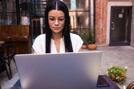 Woman skilled copywriter using applications on laptop computer, sitting in coffee shop outdoors. Female in fashionable glasses checking e-mail on notebook gadget, resting in cafe outsideの写真素材