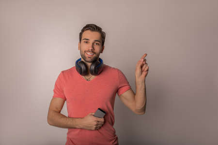 Portrait of a happy smiling hipster guy with mobile phone in hands and wireless headphones on neck showing finger on copy space for promotional content, standing isolated in studioの写真素材