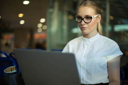 Confident woman employer in spectacles checking e-mail via laptop computer while sitting in airport terminal before business trip. Female government worker using applications on notebook gadgetの写真素材