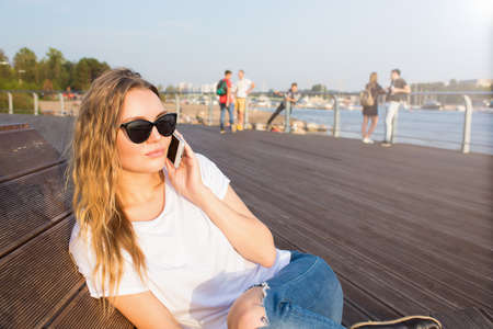 Young blonde woman talking via mobile phone while sitting on embarkment river in sunny summer evening during leisure time. College female having cell telephone conversation while relaxing outdoorsの写真素材