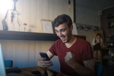 Happy smiling man freelance worker celebrating victory with winning gesture while reading on mobile phone e-mail with good news while sitting with netbook in coffee shop interiorの写真素材