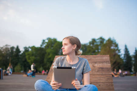 Young thoughtful female skilled content writer thinking during freelance work on web page via portable touch pad while relaxing in park in summer day. Pondering woman using digital tablet for educationの写真素材