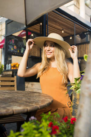 Gorgeous lady with beautiful smiling on the face in dress and wicker hat posing for camera while sitting in restaurant outdoors in sunny summer dayの写真素材