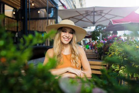 Portrait of a happy smiling charming carefree woman in elegant dress and hat with good mood smiling for camera while sitting in coffee shop outdoors during recreation time in vacation abroadの写真素材