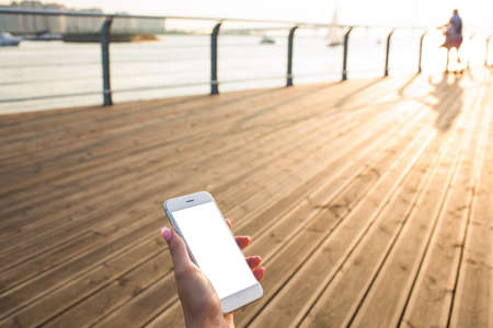 Female holding mobile phone with empty mock up copy space screen background for promotional content while relaxing outdoors on wooden embankment in sunny summer day. Cellphone with blank displayの写真素材