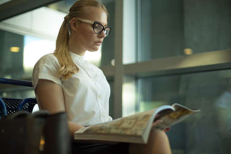 Successful woman leadership in glasses reading magazine while sitting in airport terminal before business trip abroad. Proud female university student reading fashion journalの写真素材