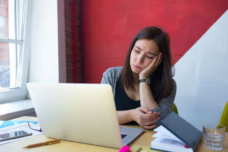Tired lazy female freelance worker reading e-mail on notebook gadget while sitting at table with...