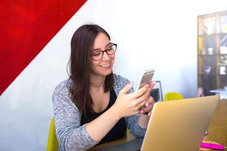 Happy smiling female in glasses skilled content writer reading e-mail on mobile phone while sitting at workplaces in co-working space. Office worker online chatting on cellphone during webinarの写真素材