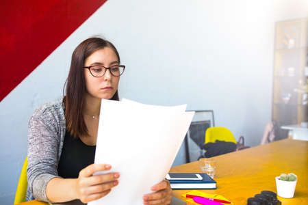Serious woman successful leadership in trendy glasses reading paper documents while sitting in co-working interior. Female financier reading summary sitting in office near copy space for advertisingの写真素材