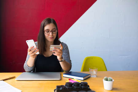 Young business woman in glasses online shopping store via mobile phone and using credit debit card for payment while sitting at workplace in office interior. Hipster girl banking via cell telephoneの写真素材