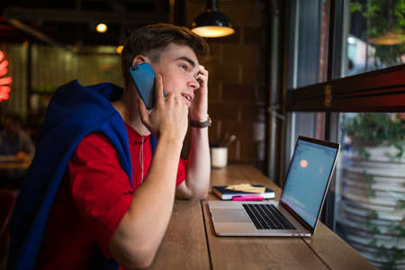 Stylish hipster guy talking via mobile phone while sitting with laptop computer in co-working cafe during free time in weekendの写真素材