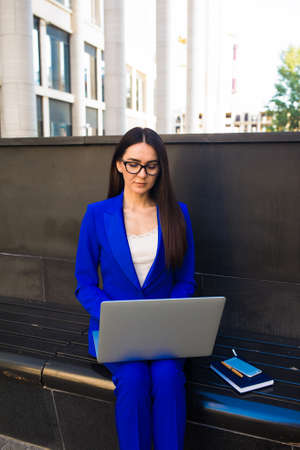 Young businesswoman in suit and fashionable glasses searching adverting information on laptop computer while sitting outdoors near companyの写真素材