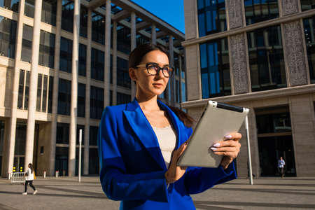 Confident female business owner in stylish spectacles and formal suit online checking e-mail on touch pad computer while standing outdoors near contemporary company buildingの写真素材