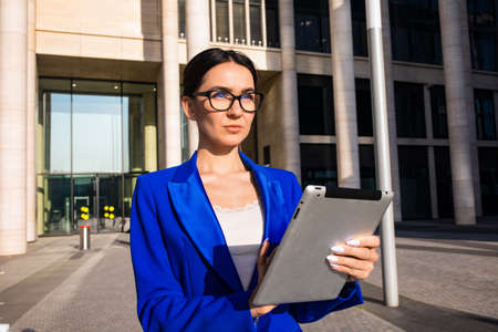 Pensive female in stylish spectacles and in suit thoughtful looking away while standing with digital...