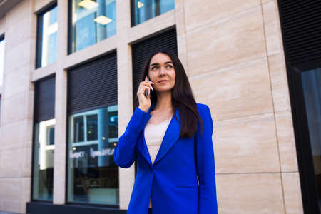 Woman in suit successful manager having cellphone conversation while standing outside modern company building during work break. Female lawyer talking via mobile phoneの写真素材