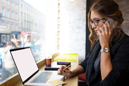 Female skilled business owner talking via mobile phone and using laptop computer with empty mock up...