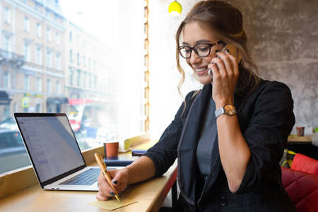 Happy smiling woman professional government worker talking via mobile phone and writing notary while sitting with laptop computer in coffee shop during work breakの写真素材