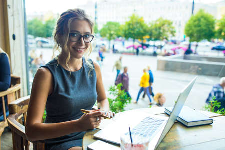 Happy smiling woman student in glasses looking in camera while sitting with pc laptop computer in coffee shop during work break. Project manager using notebook gadgetの写真素材