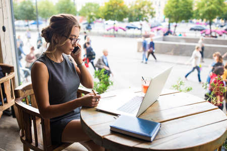 Female in glasses skilled internet marketer having cellphone conversation and writing notary while sitting with notebook gadget in restaurant during work break.の写真素材