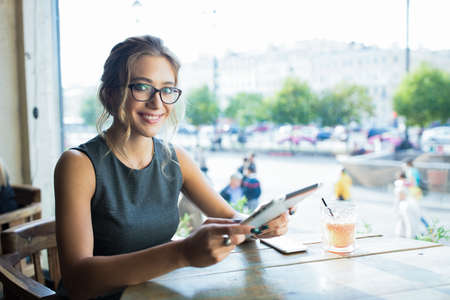 Happy smiling woman in glasses professional hostess holding in hand touch pad and looking in camera while sitting in restaurantの写真素材