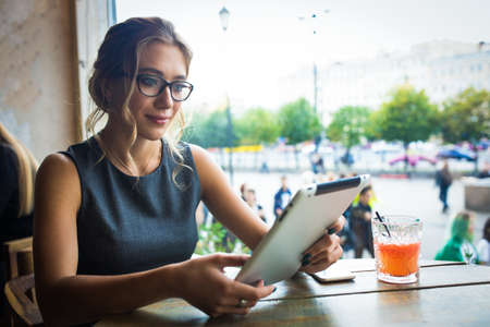 Serious woman in glasses prosperous internet marketer reading blog on website via touch pad computer while sitting in coffee shop. Female magazine editor using digital gadget for online workの写真素材