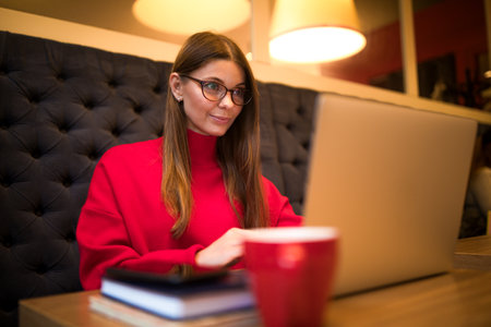 Gorgeous woman in glasses skilled marketing writer reading newsletter on web page via laptop computer while sitting in coffee shop interiorの写真素材