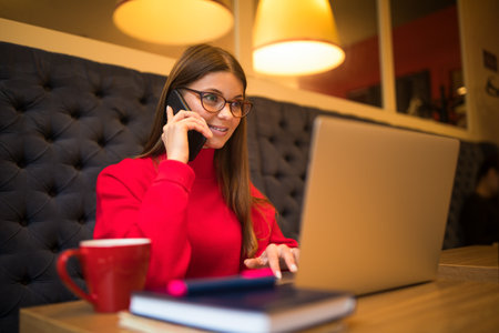 Business woman skilled marketing coordinator having smartphone conversation during online work on laptop computer while sitting in restaurant interiorの写真素材