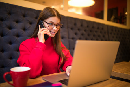 Woman in red sweater professional data conversion specialist talking via mobile phone and reading text on laptop computer while sitting in coffee shopの写真素材