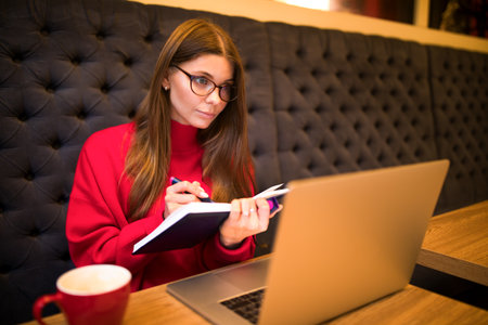 Female in glasses smart publication specialist writes in a diary and looking in laptop computer screen during online lessons while sitting with cup of coffee in restaurant interiorの写真素材
