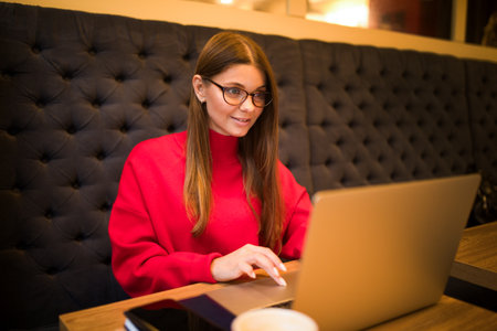 Young female in glasses and warm red sweater professional resume writer checking e-mail via laptop computer while sitting in coffee shop during free time in autumn dayの写真素材