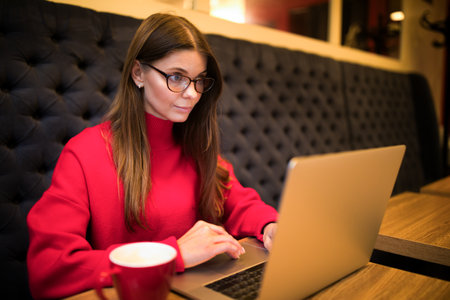 Woman in glasses and warm red sweater skilled social media manager reading notifications on laptop computer while sitting in coffee shop during work breakの写真素材
