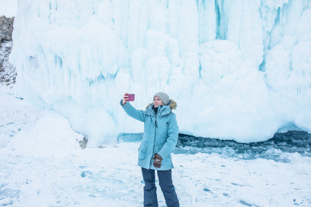 Happy smiling female traveler wearing a hat and warm clothes takes a selfie or talking on a video call via smartphone while standing against big ice on the rocksの写真素材