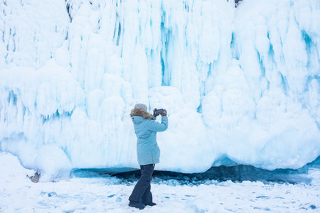 Woman in warm thermal clothing stands with back and takes a photo on a mobile phone of a big ice on the rocks mountainの写真素材
