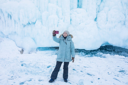 Happy smiling female traveler taking a selfie on mobile phone while standing against a large icy rock in cold winter day on frozen Baikal lakeの写真素材