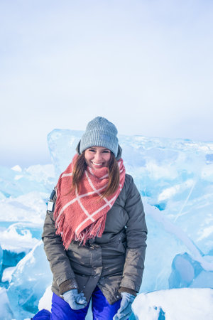 Smiling joyful female traveler with good mood looking in camera while sitting on frozen Baikal lake against big against big ice floes in winter dayの写真素材