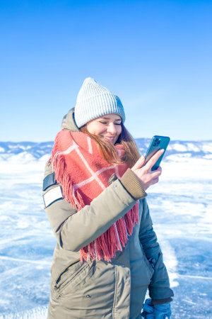Happy smiling woman traveler online chatting on mobile phone while standing outdoors on frozen Baikal lake in sunny winter dayの写真素材
