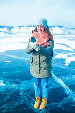 Happy smiling woman traveler online chatting on mobile phone while standing outdoors on frozen Baikal lake in winter dayの写真素材
