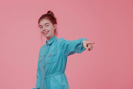 Young woman in a turquoise sweater pointing to the side with an enthusiastic smile against a pink backdropの素材