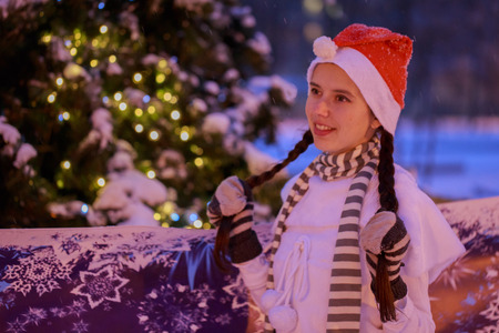 Night. A smiling girl in a red cap standing at the Christmas tree. She holds pigtails.の写真素材
