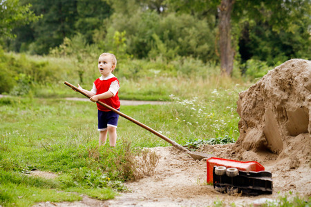 Countryside. Little boy holding a big shovel in his hands. He plays in the sand, to imitate adults.の写真素材