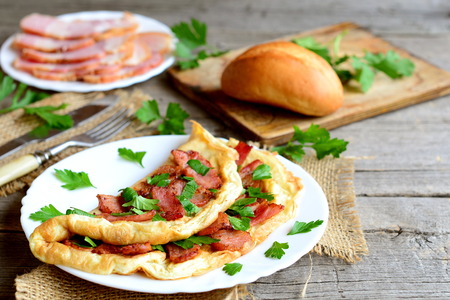 Homemade bacon omelette. Delicious omelette stuffed with bacon and parsley on a plate, bacon slices on a plate, bread roll, fork, knife, cutting board, fresh parsley sprigs on old wooden backgroundの写真素材
