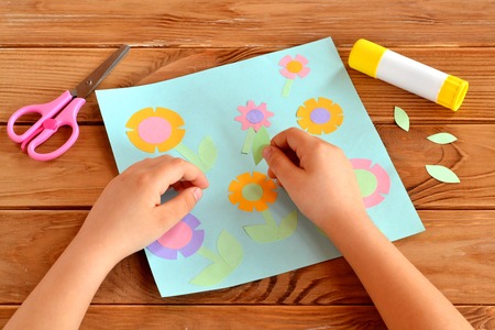 Child doing a card with flowers. Child holds a paper leaf in his hands and sticks it. Glue, scissors, greeting card on a wooden table. Paper flower craft for kids. Child art projectの写真素材
