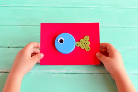 Fish crafts. Child holding a cardboard with craft fish. DIY bottle caps. Creative bottle cap ideas....