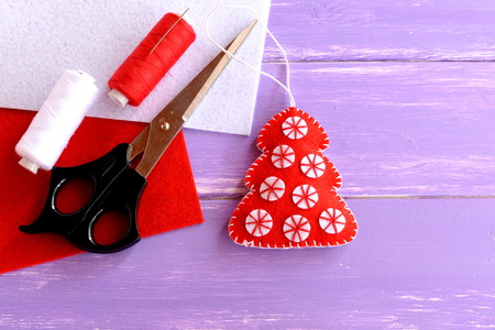 Red felt Christmas tree ornament, scissors, red and white felt sheets, thread, needle lilac on wooden background with blank space for text. Home decor. Crafts idea for Christmasの写真素材