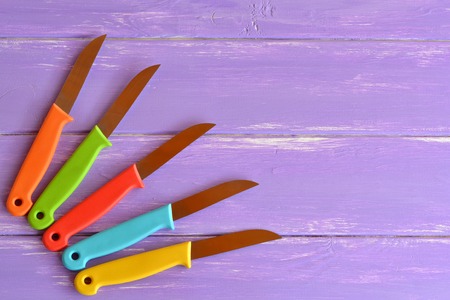 Kitchen knife set. Bright color handle. Knives with metal blade and plastic handles. Different tools for cutting. Purple wooden background with blank space for text. Top viewの写真素材