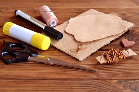 Cardboard box piece, torn wrapping paper, brown wax cord, scissors, black marker, glue stick, thread, needle, button heart, cord bow. Set to create greeting card. Kids crafts set. Wooden backgroundの写真素材