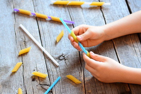 Development of fine motor skills for kids. Small kid holds a straw and a raw pasta in his hands. Creative kids activity. Easy exercise to improve hand and eye coordination. Wooden backgroundの写真素材