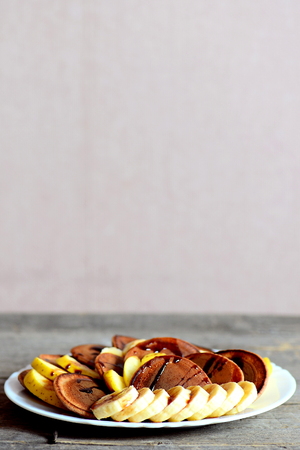 Healthy cacao pancakes with bananas, apples and caramel sauce on a white plate isolated on wooden background. Copy space for text. Pancake day breakfast and brunch recipe. Vertical photoの写真素材