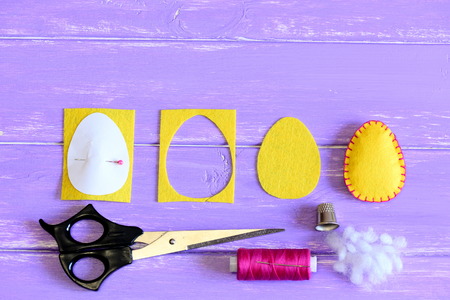 How to make a felt Easter egg ornament. Yellow felt Easter egg, cut felt piece in shape of a egg, paper template, scrap, scissors, thread, thimble on a wooden background with copy space for textの写真素材