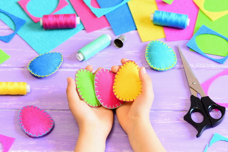 Child holding a felt Easter eggs in his hands. Little child made Easter crafts of felt. Tools and materials for needlework on wooden table. Simple kids crafts conceptの写真素材